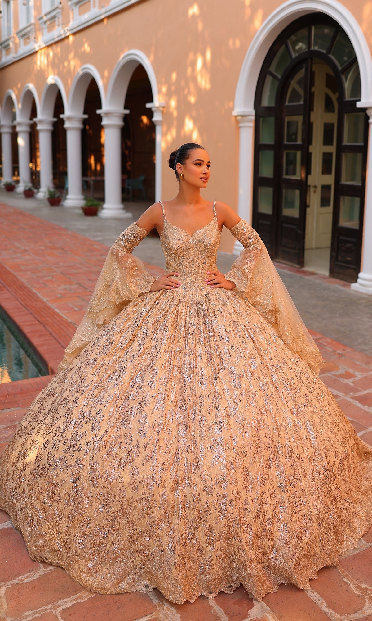 A woman stands outside a building wearing the Quinceanera Dress 54303 By Amarra: an ornate, gold, floor-length v-neck ball gown with flared sleeves.